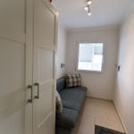 Ein kleiner Raum mit grauem Sofa, weißen Wänden und einem Fenster mit Jalousie.