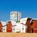 Strandkörbe auf Sandstrand mit Hochhaus im Hintergrund.
