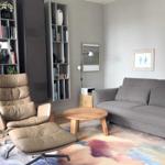 Wohnzimmer mit grauem Sofa, braunem Sessel und runden Tisch auf buntem Teppich.