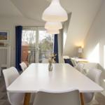 Essbereich mit weißem Tisch und Stühlen. Hintergrund: Wohnzimmer mit Balkon und Fenster.