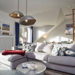 Wohnzimmer mit grauem Sofa, blauen Vorhängen und Holzboden. An der Wand hängt ein Bild.