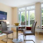 Essbereich mit Glasrundtisch, drei Stühlen und TV an der Wand. Blick auf grünen Garten.