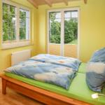 Schlafzimmer mit Holzbett, grünem Matratzenbezug und Fenstern mit Blick auf Grün.