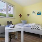 Ein Schlafzimmer mit Bett, Tisch und Stühlen. An der Wand hängen Bilder. Durch das Fenster ist ein Balkon sichtbar.