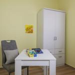 Ein Raum mit weißem Schreibtisch, Stuhl und Kleiderschrank an der Wand.