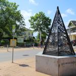 Draußen steht eine schwarz lackierte Pyramide auf Steinsockel mit Inschrift. Hintergrund sind Gebäude und Bäume.