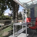 Terrasse mit Tisch, zwei roten Stühlen und Glasgeländer. Blick auf grüne Landschaft und Gebäude.
