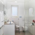 Badezimmer mit Dusche, Waschbecken und Toilette. Weiße Fliesen und Fenster sorgen für viel Licht.