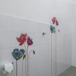 Dusche mit weißen Fliesen und bunten Blumenmotiven an der Wand. Toilettenpapierhalter sichtbar.