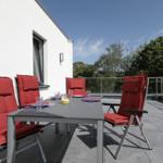 Terrasse mit Tisch und vier Stühlen mit roten Sitzkissen. Hintergrund: weißes Haus und Bäume.
