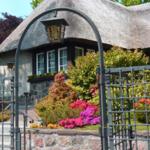 Haus mit Strohdach, Steinmauer und Garten mit Blumen hinter Eisentor.