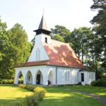 Weiße Kirche mit rotem Dach und spitzen Turm in grüner Umgebung.