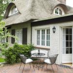 Haus mit Strohdach, Terrasse mit Tisch und Stühlen, Fenster und Glastüren.