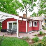 Rotes Holzhaus mit Terrasse und Sitzgelegenheit unter Glasdach. Umgeben von Grün und Bäumen.