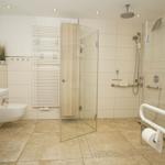 Badezimmer mit Dusche, WC und Waschbecken. Fliesenboden und Handlauf für barrierefreien Zugang.
