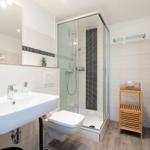 Badezimmer mit Dusche, Waschbecken und Toilette. Holzregal und Heizkörper.