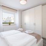 Schlafzimmer mit Doppelbett, weißen Schränken und Fensterblick auf Wohnhaus.