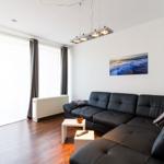 Wohnzimmer mit schwarzer Ledercouch, weißen Tischen und Holzboden. An der Wand hängt ein Bild.