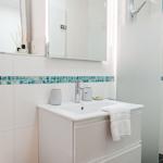 Bad mit Waschbecken, Spiegel und Dusche. Wandfliesen mit blauem Mosaikstreifen.