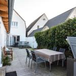 Hinterhof mit Holzterrasse, Tisch, Stühlen und Beerenhecke. Hintergrund: weiße Häuser mit Dächern.