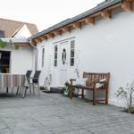 Terrasse mit Holzbank, Tisch und Stühlen. Weiße Wand, Fenster und Tür.