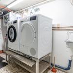 Wasch- und Trockner stehen auf Metallgestell. An der Wand hängt ein Waschbecken mit Armatur.