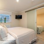 Schlafzimmer mit Bett, TV und Fenster. Durch Glastür ist zweites Schlafzimmer sichtbar.