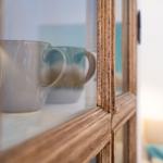 Eine Tasse steht in einem Holzregal mit Glasfront. Im Hintergrund ist ein Sofa mit Kissen sichtbar.