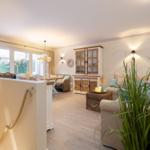 Wohnbereich mit Sofa, Tisch und Holzschränken. Große Fenster und Pflanzen sorgen für viel Licht.