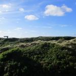 Weitläufige Dünenlandschaft unter blauem Himmel mit vereinzelten Wolken und einem Holzgestell im Hintergrund.