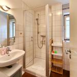 Badezimmer mit Dusche, Waschbecken und Spiegel. Holzregal und Fenster sichtbar.