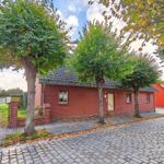 Rotes Backsteingebäude mit Eingangstür und Fenstern unter Bäumen auf Kiesweg.