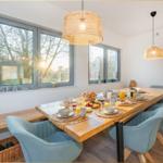 Esszimmer mit langem Holztisch, blauen Stühlen und großen Fenstern mit Blick auf Garten.