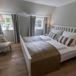 Ein Schlafzimmer mit weißem Bett, Holzboden und zwei Fenstern mit Vorhängen.