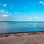 Strand mit Steg und Badegästen im klaren Meer unter blauem Himmel.