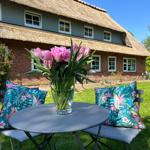 Runder Tisch mit Blumen und Kissen im Garten vor einem Strohdachhaus.