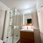 Badezimmer mit Dusche, Waschbecken und Toilette. Fliesenwand und beleuchtetes Spiegel.