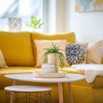 Gelber Sofa mit Kissen und Pflanzen auf runden Tischen.