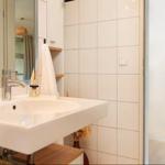 Waschbecken mit Spiegel, weiße Fliesen und Holzregal. Duschbereich mit Glaswand.