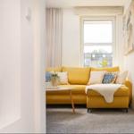 Ein gelbes Sofa mit Kissen und Decke steht vor einem Fenster. Links ist eine Glastür zu sehen.