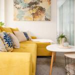 Gelber Sofa mit Kissen, runder Tisch und Wandbild mit Bäumen.