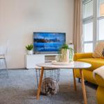 Ein Wohnzimmer mit gelbem Sofa, Fernseher und Esstisch. Pflanzen und Lampen sind vorhanden.