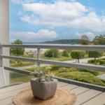 Balkon mit Tisch und Pflanze. Blick auf Grünflächen, Straße und Wasser.