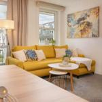 Wohnzimmer mit gelbem Sofa, Tisch und Fenstern. Schlafzimmer im Hintergrund.