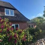 Haus mit Ziegelfassade und Dachgauben, umgeben von Blumen und Bäumen unter blauem Himmel.