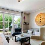 Wohnzimmer mit Sofa, Sessel und Tisch. Große Fensterfront mit Blick auf Garten.