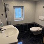 Badezimmer mit Waschbecken, Toilette und Fenster. Fliesen und Spiegel sichtbar.
