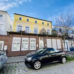 Gelbes Haus mit Balkon und Klinkerfassade. Autos parken auf Kopfsteinpflaster.