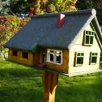 A small wooden house with a gray roof and green window frames stands on a post in the garden.