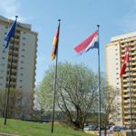 Zwei Hochhäuser mit Flaggen und Bäumen im Vordergrund. Parkplatz mit Autos.